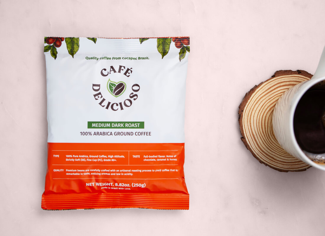A package of Café Delicioso coffee next to a cup on a wooden coaster, against a light pink backdrop.