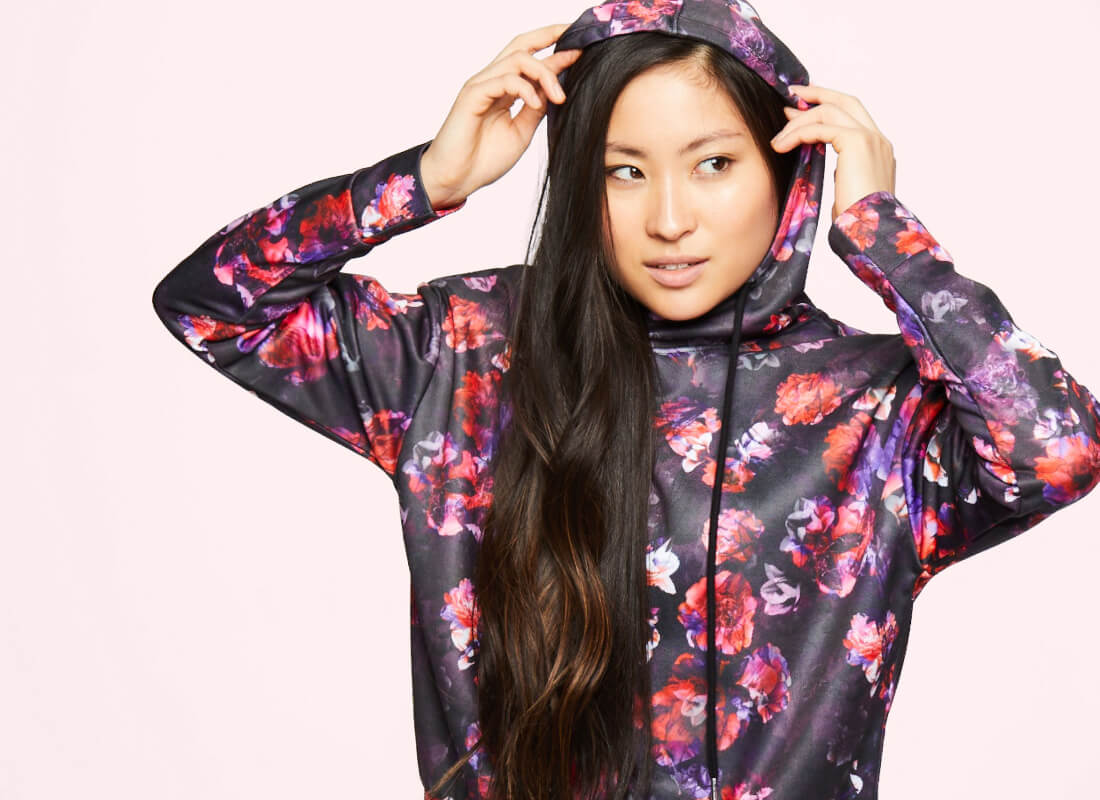 A woman in a colorful floral-print hoodie poses against a pink background, playfully adjusting her hood. Long, dark hair cascades down.