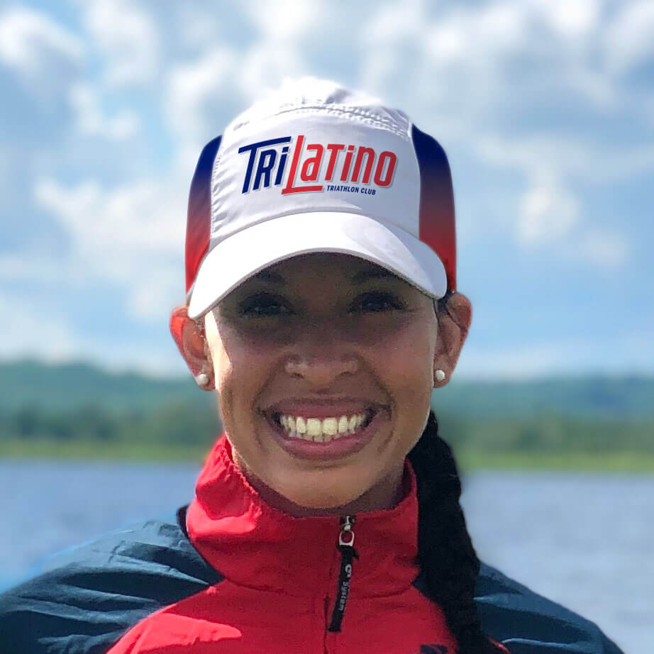 A woman wearing a red and blue cap with "TriLatino Triathlon Club" logo, dressed in a red jacket, stands by a lake under a cloudy sky.