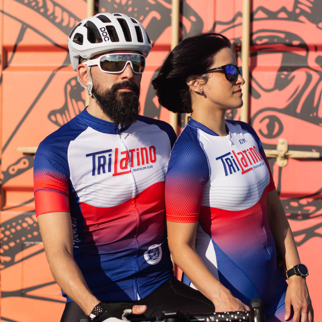 A man and woman pose back-to-back, wearing matching blue, red, and white jerseys of the TriLatino Triathlon Club against a colorful background.