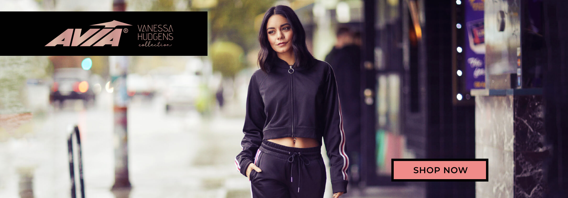 Vanessa Hudgens models a stylish black athleisure outfit with white stripes on a rainy street, promoting the AVIA Vanessa Hudgens collection with its logo and “shop now” button.