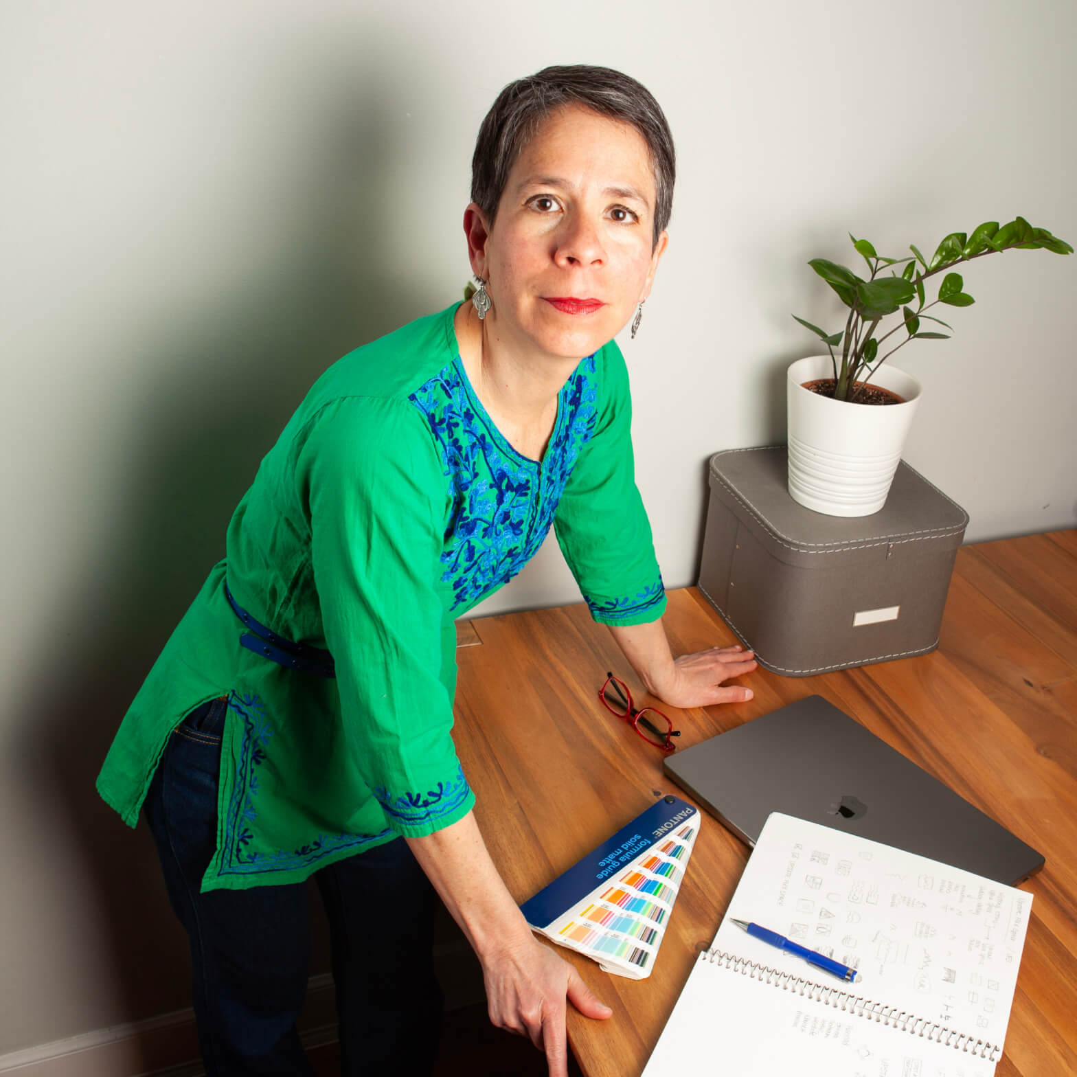 A woman, Veronica Choroco in a bright green embroidered top, leans over a wooden table with color swatch, a laptop, notebook and a plant in view.