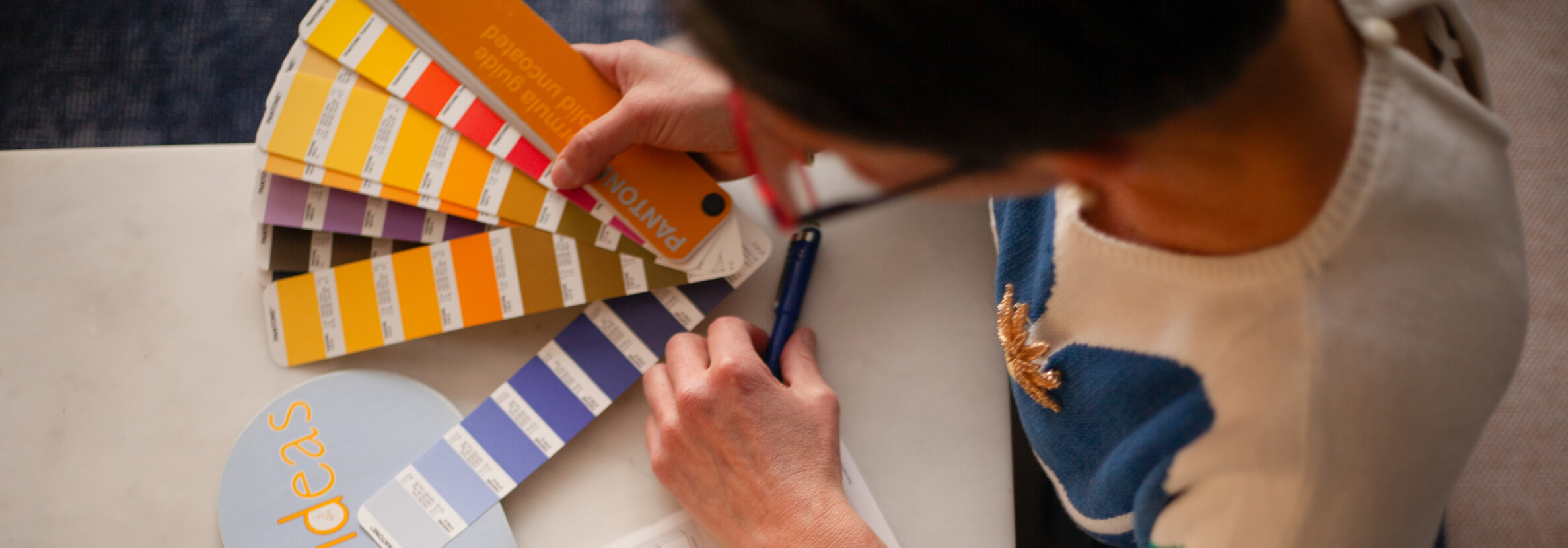 A woman, Veronica Choroco, holds color swatches, analyzing and marking selections for a design project on a white table.