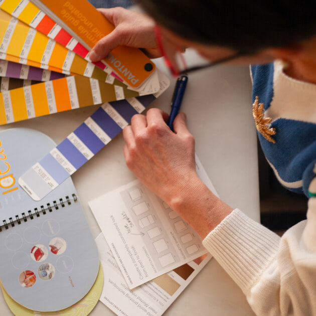 A woman, Veronica Choroco, holds color swatches, analyzing and marking selections for a design project on a white table.