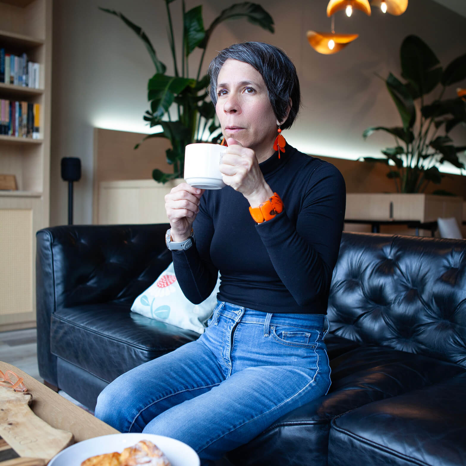 A woman, Veronica Choroco, is sitting on a couch, holding a white mug, wearing a black top and blue jeans, with a plate of croissants nearby and plants in the background.